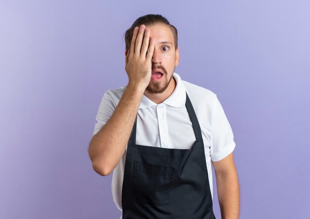 Impressed young handsome barber wearing uniform closing half of his face with hand isolated on purple  with copy space