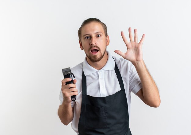 Impressed young handsome barber wearing uniform holding hair clippers and showing five with hand isolated on white  with copy space