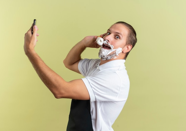 Впечатленный молодой красивый парикмахер в униформе держит мобильный телефон и наносит крем для бритья на собственную бороду, изолированную на оливково-зеленом цвете с копией пространства