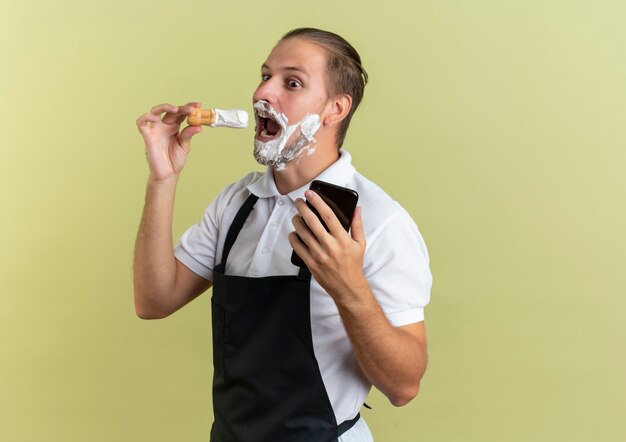 Impressed young handsome barber wearing uniform holding mobile phone and trying to put shaving brush into his mouth looking at side isolated on olive green  with copy space