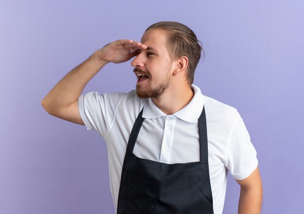 Impressed young handsome barber wearing uniform keeping hand above eyes looking at distance isolated on purple 