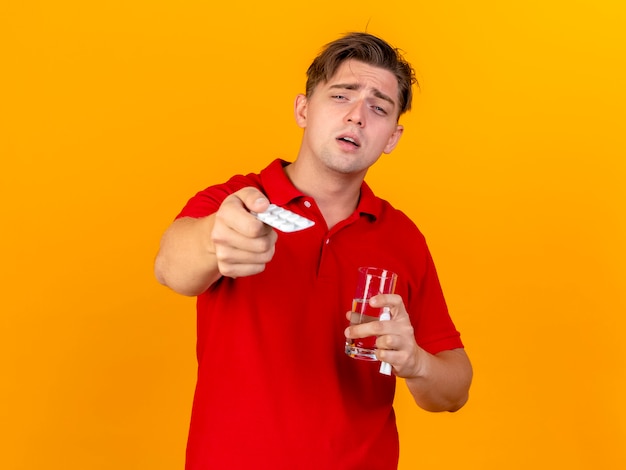 Impressed young handsome blonde ill man holding pack of medical tablets glass of water and napkin