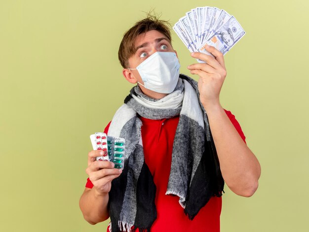 Impressed young handsome blonde ill man wearing mask holding money and packs of medical pills looking at money isolated on olive green wall