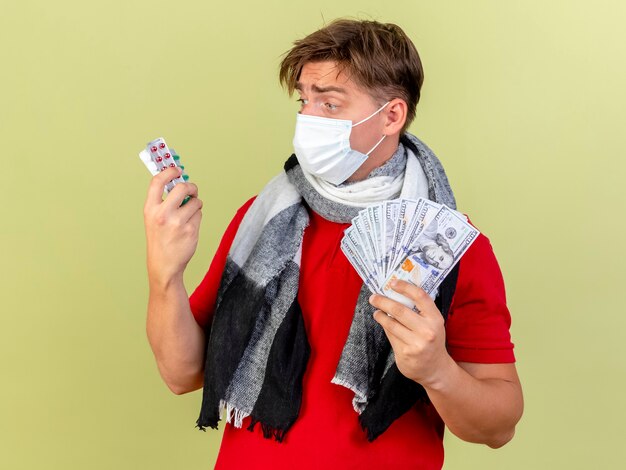 Impressionato giovane uomo malato biondo bello che indossa maschera e sciarpa tenendo i soldi e le pillole guardando le pillole isolate su sfondo verde oliva