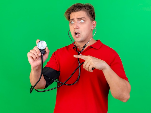 Впечатленный молодой красивый блондин больной мужчина со стетоскопом, измеряющим давление самому себе, смотрит и указывает на сфигмоманометр, изолированный на зеленой стене