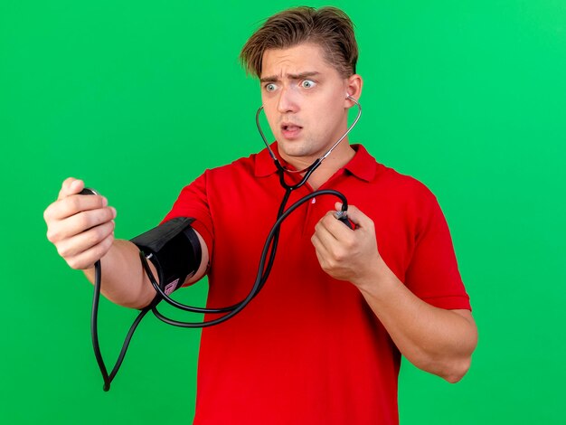 Impressed young handsome blonde ill man wearing stethoscope measuring pressure to himself holding and looking at sphygmomanometer isolated on green wall