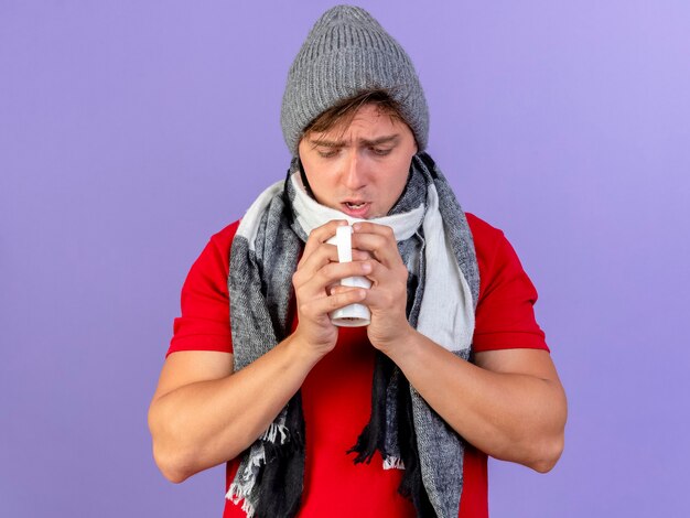 Impressed young handsome blonde ill man wearing winter hat and scarf holding and looking at cup of tea isolated on purple wall