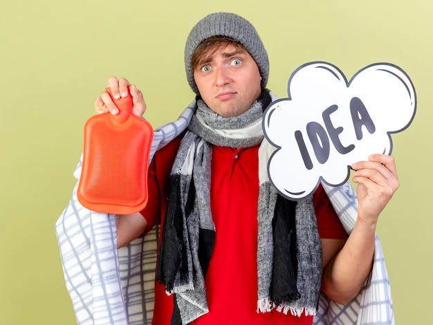 Free photo impressed young handsome blonde ill man wearing winter hat and scarf wrapped in plaid holding idea bubble and hot water bottle looking at camera isolated on olive green background