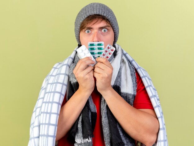 Free photo impressed young handsome blonde ill man wearing winter hat and scarf wrapped in plaid touching mouth with packs of medical pills looking at camera isolated on olive green background