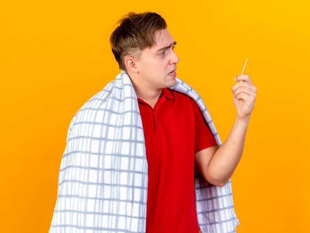 Impressed young handsome blonde ill man wrapped in plaid holding and looking at thermometer isolated on orange wall