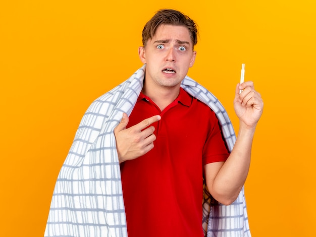 Impressed young handsome blonde ill man wrapped in plaid holding and pointing at thermometer isolated on orange wall