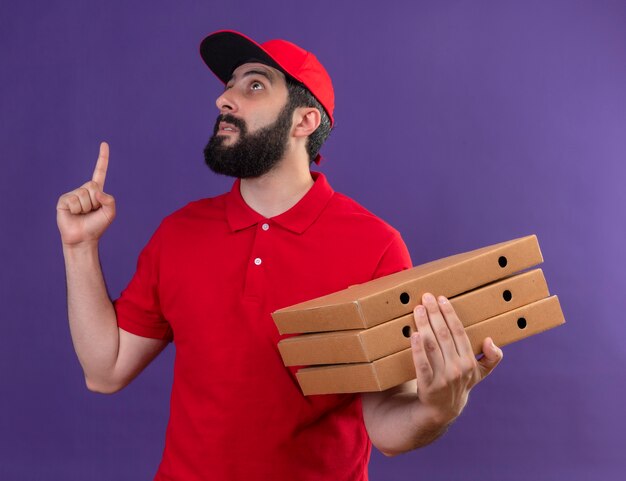 Impressed young handsome caucasian delivery man wearing red uniform and cap holding pizza boxes looking and pointing up isolated on purple 