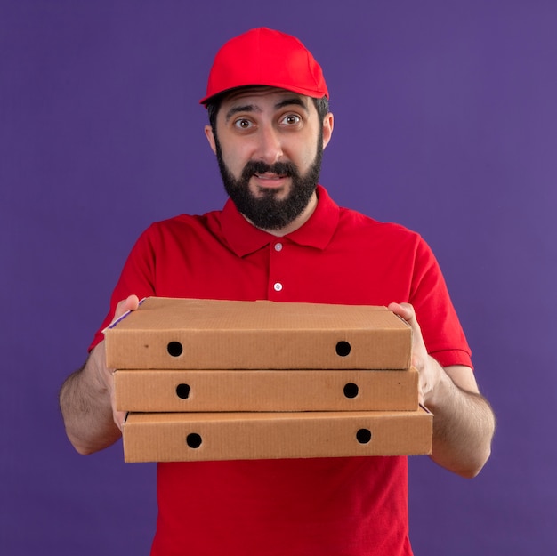 빨간색 유니폼과 보라색에 고립 된 피자 상자를 뻗어 모자를 입고 감동 젊은 잘 생긴 백인 배달 남자