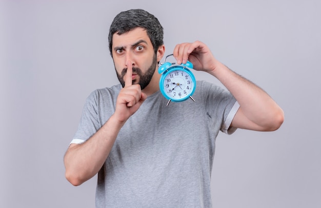 Impressed young handsome caucasian man holding alarm clock and gesturing silence isolated on white 