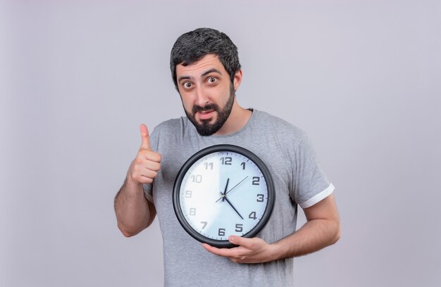 Impressed young handsome caucasian man holding clock and showing thumb up isolated on white  with copy space