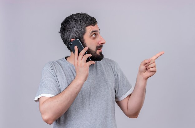 Impressed young handsome caucasian man talking on phone looking and pointing at side isolated on white 