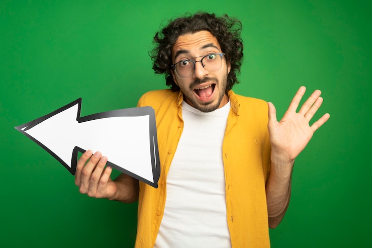 मुफ़्त फोटो impressed young handsome caucasian man wearing glasses holding arrow mark that is pointing at side  showing empty hand isolated on green wall