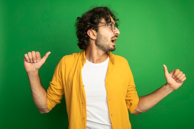 Impressed young handsome caucasian man wearing glasses pointing at himself looking at side isolated on green background