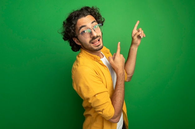 Impressed young handsome caucasian man wearing glasses standing in profile view looking at camera pointing up isolated on green background with copy space