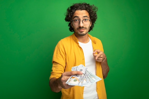 Free photo impressed young handsome caucasian man wearing glasses stretching out money  pointing at it  isolated on green wall with copy space