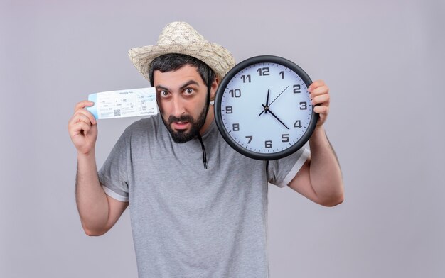 Impressed young handsome caucasian traveler man wearing hat holding clock and airplane ticket isolated on white  with copy space