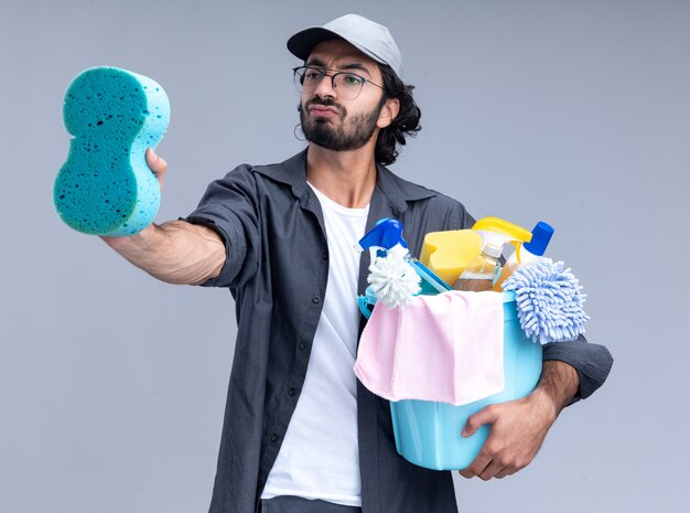 Impressed young handsome cleaning guy wearing t-shirt and cap holding bucket of cleaning tools and looking at sponge in his hand isolated on white wall