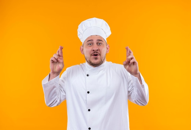 Impressionato giovane bel cuoco in uniforme da chef che fa un gesto di dita incrociate isolato sul muro arancione orange