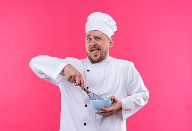Impressed young handsome cook in chef uniform holding bowl and whisk isolated on pink wall