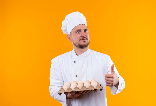 오렌지 벽에 엄지 손가락을 보여주는 계란의 상자를 들고 요리사 유니폼에 감동 젊은 잘 생긴 요리사