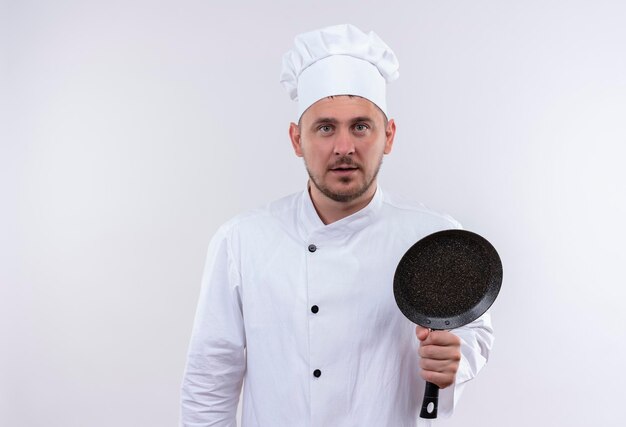 Impressed young handsome cook in chef uniform holding frying pan isolated on white wall
