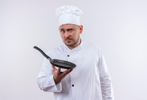 Impressed young handsome cook in chef uniform holding frying pan isolated on white wall