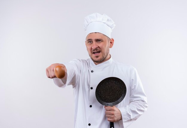 Impressed young handsome cook in chef uniform holding frying pan and pointing with spoon isolated on white wall