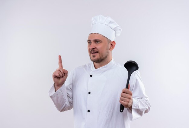 鍋を持ち、白い壁に孤立した側を見て指を上げるシェフの制服を着た若いハンサムな料理人に感銘