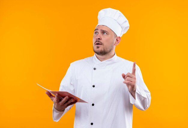 シェフの制服を着た印象的な若いハンサムな料理人がメモ帳を持ち、オレンジ色の壁に孤立した見上げる指を上げる
