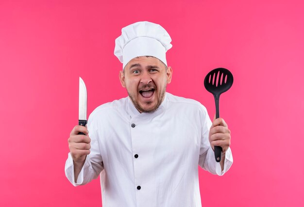 Impressed young handsome cook in chef uniform holding slotted spoon and knife isolated on pink wall