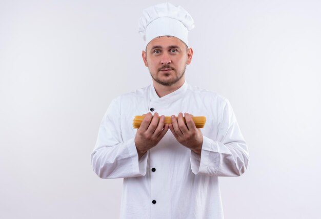 Impressed young handsome cook in chef uniform holding spaghetti pasta isolated on white wall