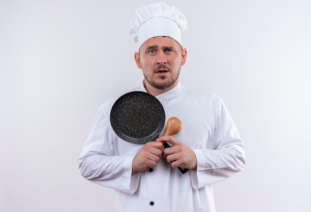 Impressed young handsome cook in chef uniform holding spoon and frying pan on isolated white wall