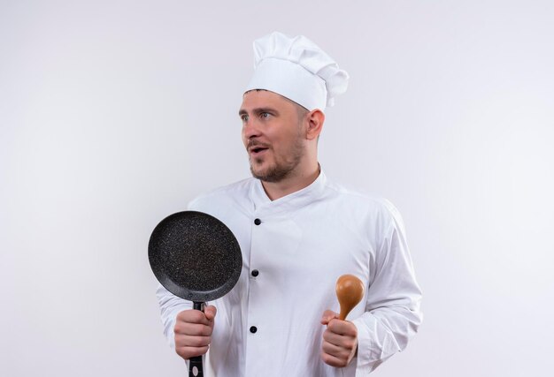 Impressed young handsome cook in chef uniform holding spoon and frying pan looking at side isolated on white wall