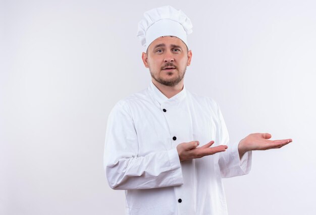 Impressed young handsome cook in chef uniform pointing with hands at side isolated on white wall