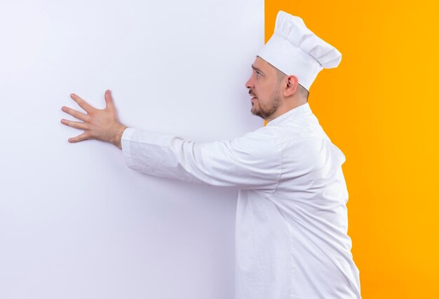 白い壁の前に立つシェフの制服を着た若いハンサムな料理人が、オレンジ色の壁に隔離された側を見て、それに手を置いた