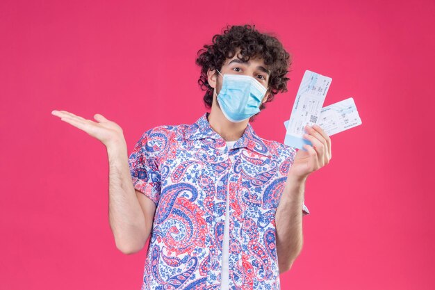 impressed young handsome curly traveler man wearing medical mask holding airplane tickets showing empty hand on isolated pink space