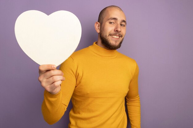 Impressed young handsome guy holding heart shape box isolated on purple