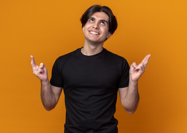 Impressed young handsome guy wearing black t-shirt crossing fingers isolated on orange wall