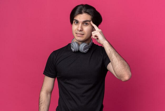 Impressed young handsome guy wearing black t-shirt and headphones around neck putting finger on temple isolated on pink wall