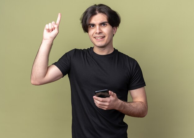 Impressed young handsome guy wearing black t-shirt holding phone points at up isolated on olive green wall