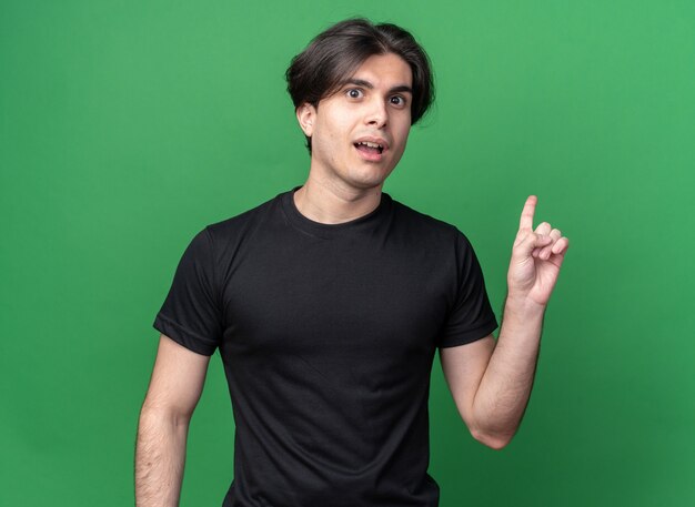 Впечатленный молодой красивый парень в черной футболке указывает на спину, изолированную на зеленой стене с копией пространства