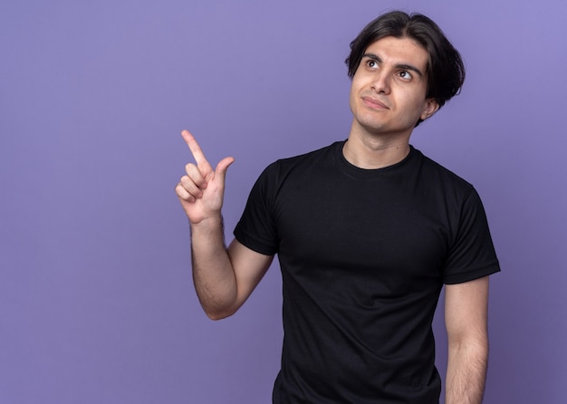 Impressed young handsome guy wearing black t-shirt points at side isolated on purple wall with copy space