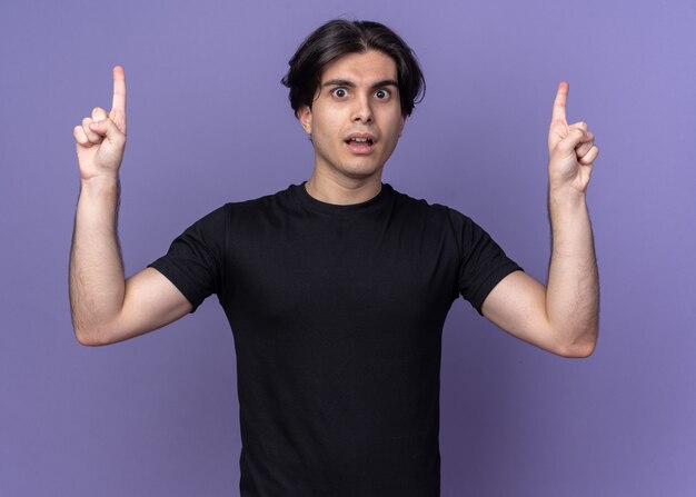 Впечатленный молодой красивый парень в черной футболке указывает вверх изолированным на фиолетовой стене