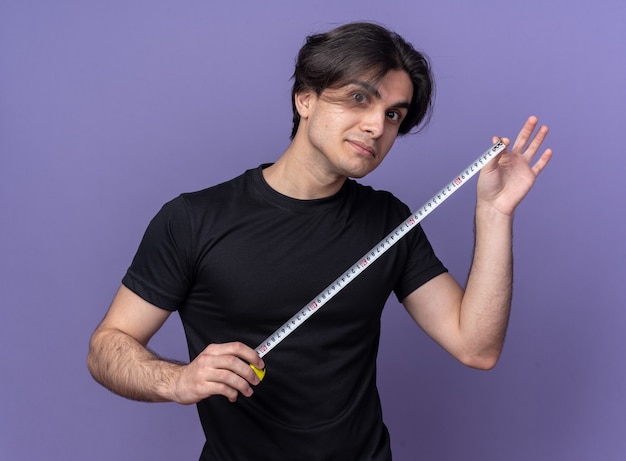 Impressed young handsome guy wearing black t-shirt stretching out tape measure isolated on purple wall