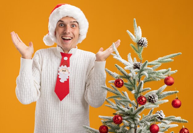Бесплатное фото Впечатленный молодой красивый парень в новогодней шапке и галстуке санта-клауса, стоящий возле украшенной елки, глядя в камеру, показывая пустые руки, изолированные на оранжевом фоне
